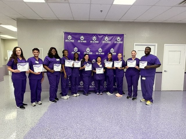 Eleven graduates in purple scrubs holding certificates beside the All is Well banner