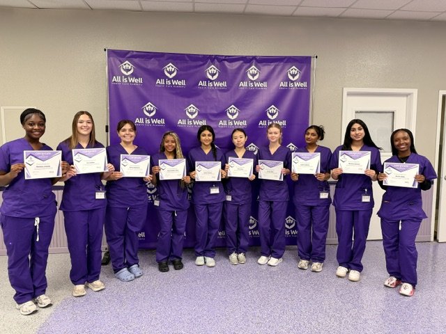 Ten graduates in purple scrubs holding certificates beside the All is Well banner